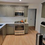 Modernized kitchen with green grey cabinetry, stainless steel appliances, light hardwood floors and white counter tops