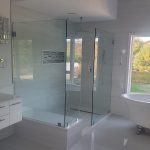 Bright and modern bathroom with rain shower head, glass shower enclosure, glossy white tiling and backsplash, matte white modern cabinetry and white cast iron tub in front of large bay windows overlooking scenic foliage