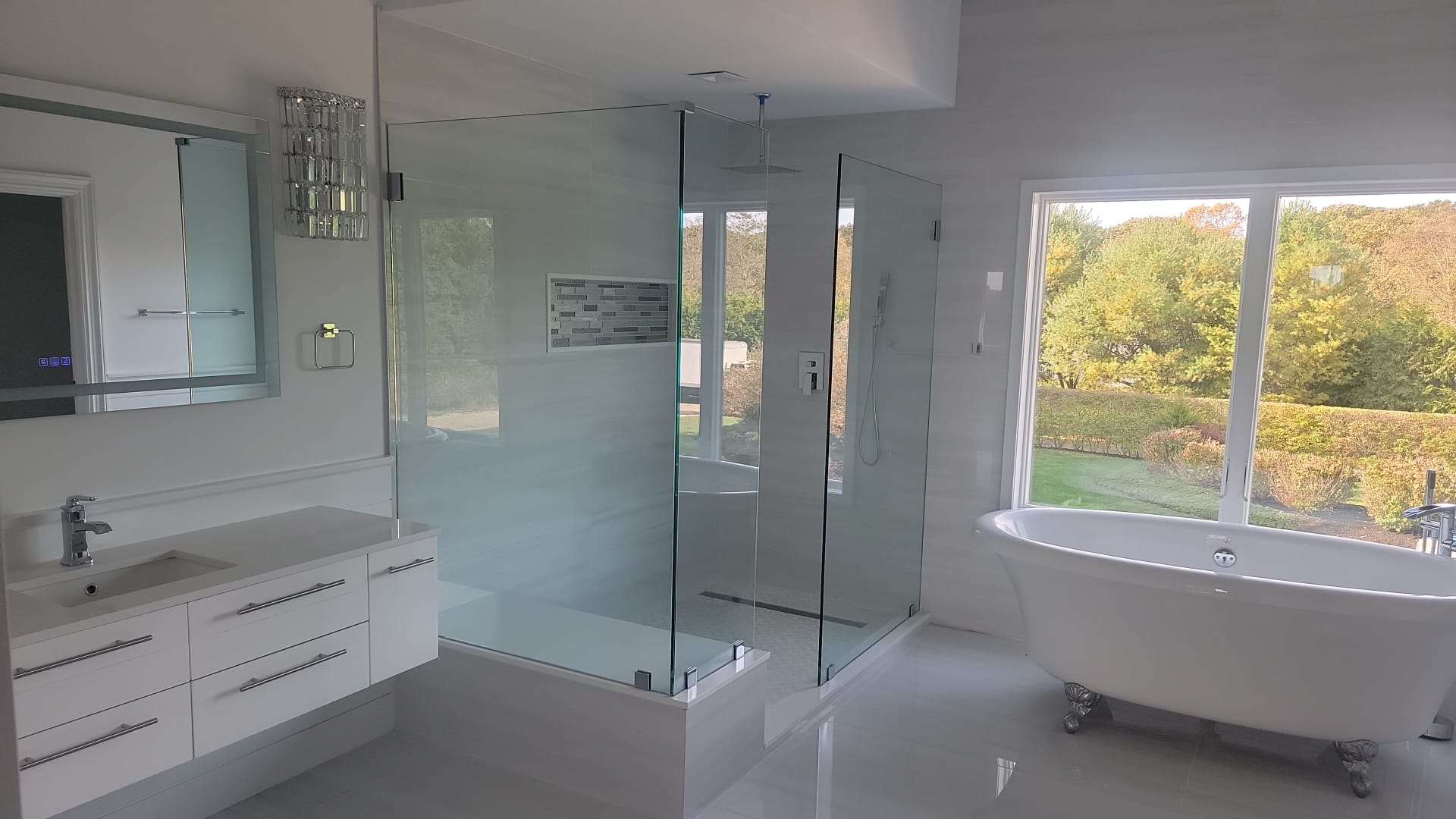 Bright and modern bathroom with rain shower head, glass shower enclosure, glossy white tiling and backsplash, matte white modern cabinetry and white cast iron tub in front of large bay windows overlooking scenic foliage